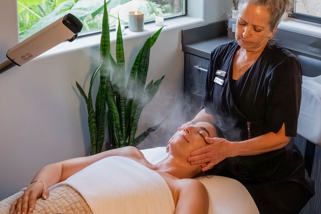A woman performs a facial treatment on a reclining client in a spa room, using gentle hands while mist rises from the treatment area, relaxing.
