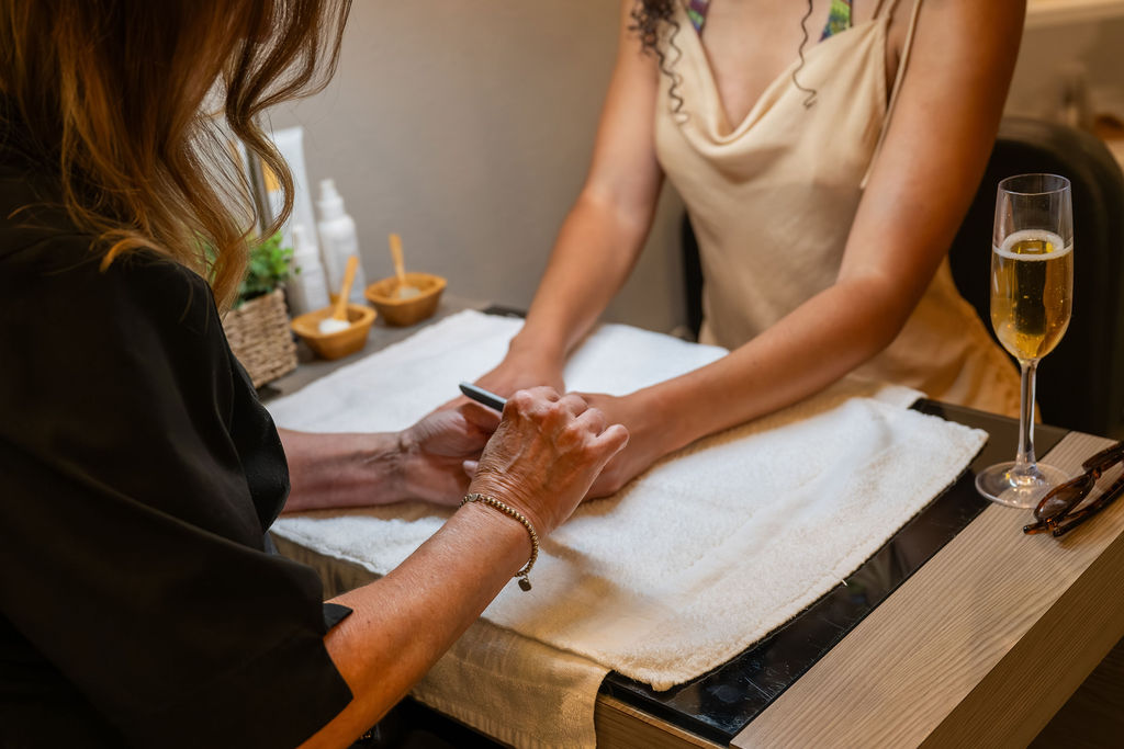 A spa massage session is underway, with a therapist gently stretching a client&rsquo;s hands while a champagne flute sits nearby, smiling.