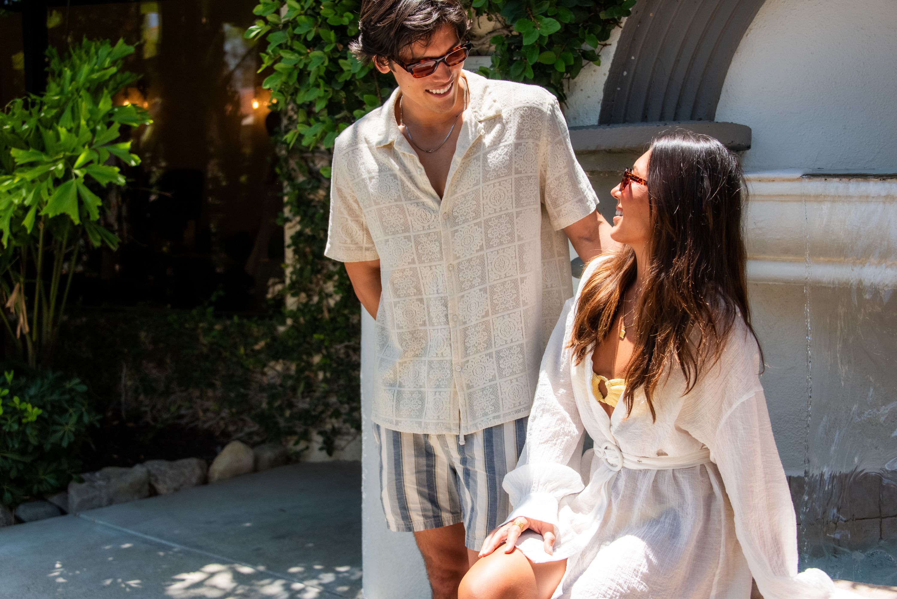 A couple wearing sunglasses in white outfits smiles together outdoors beside lush green plants, enjoying sunny patio vibes.