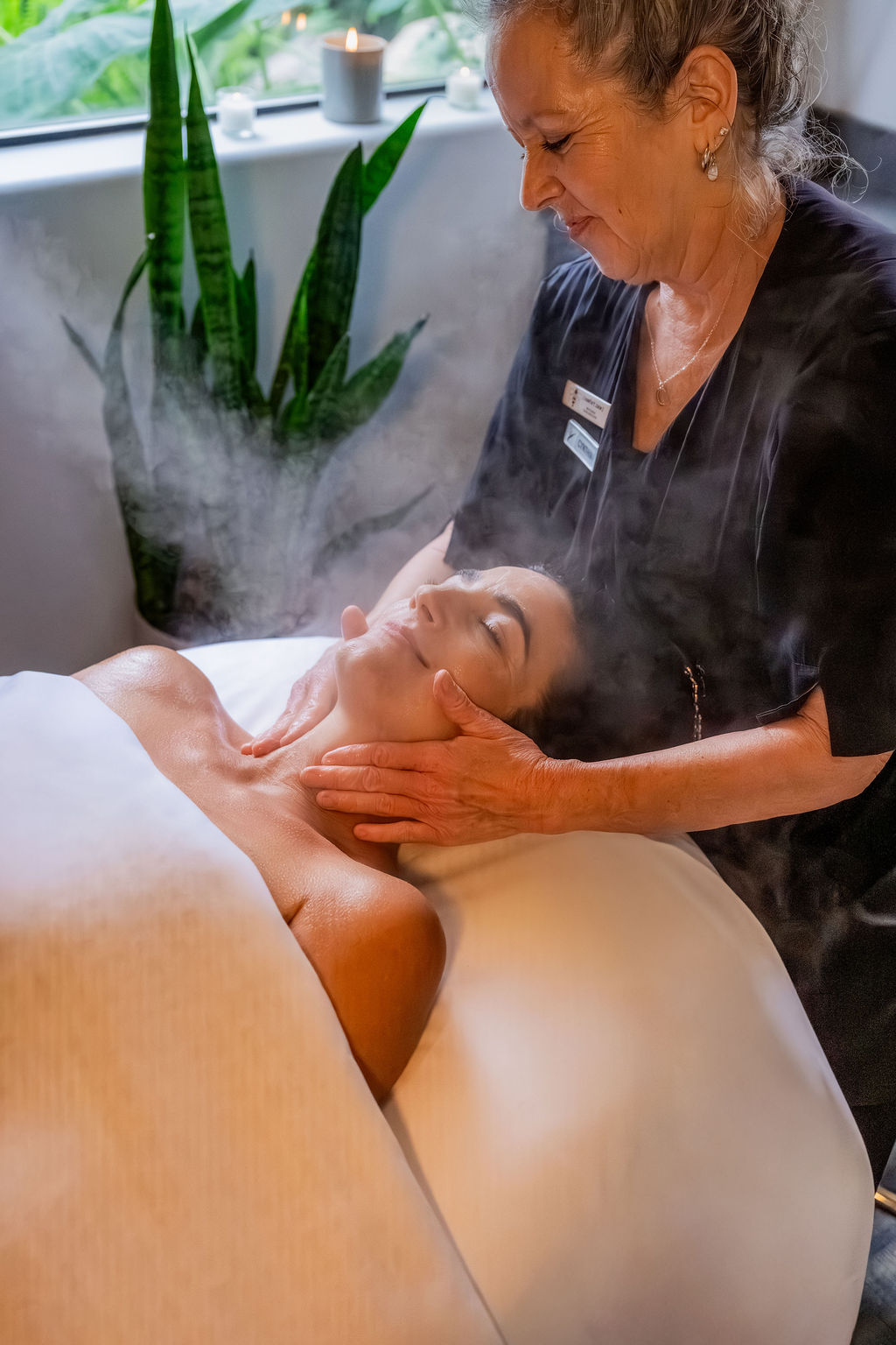 An elderly therapist performs a facial massage with steam in a spa, gently supporting a relaxed young client&rsquo;s face, shoulders, and neck.
