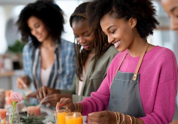 A group of women in aprons in a kitchen prepare drinks and mix ingredients at a counter, smiling while working together.
