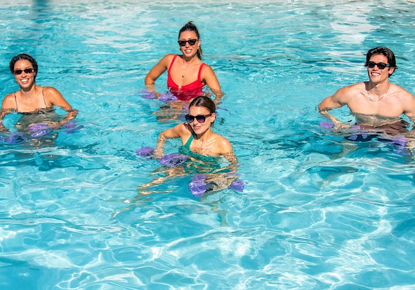 Four people in swimwear float and smile in a clear blue swimming pool, wearing sunglasses and posing together in the water.