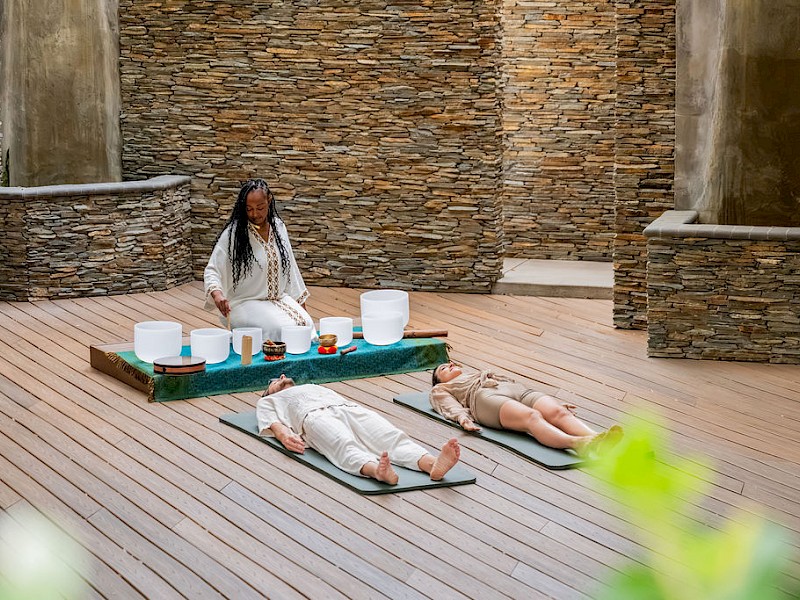 A woman leads a yoga/meditation session in a stone-walled studio, with two people lying on mats and candles on a table.