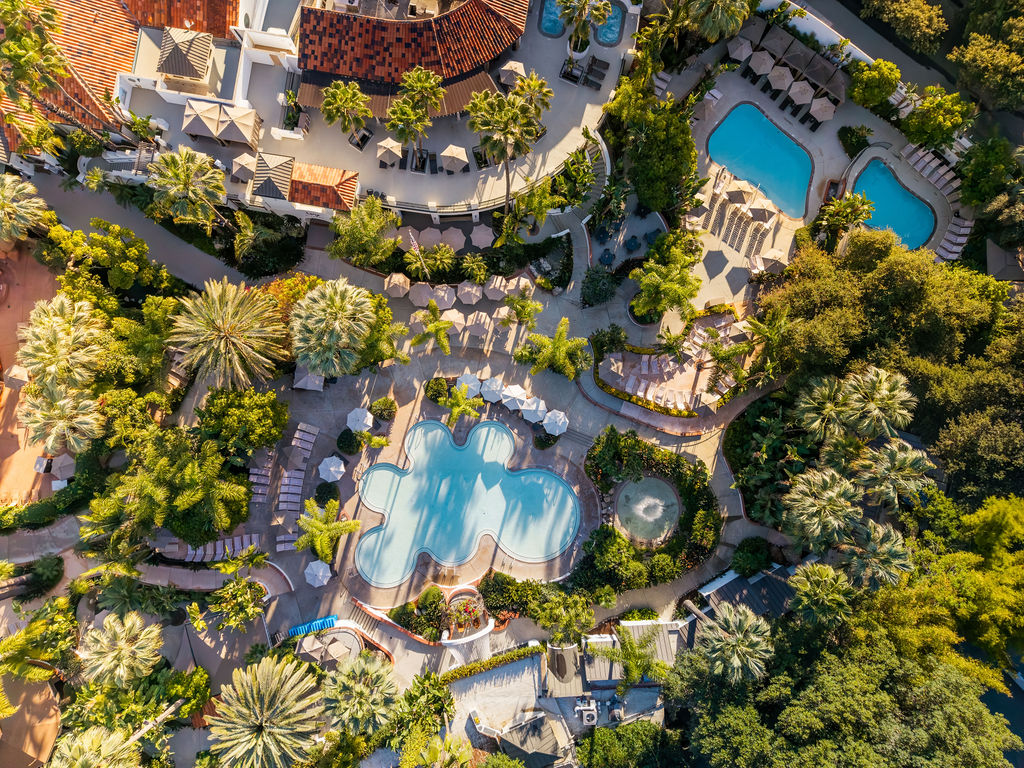 Aerial view of a tropical resort with multiple swimming pools, palm trees, and landscaped pathways, all under bright sunlight.
