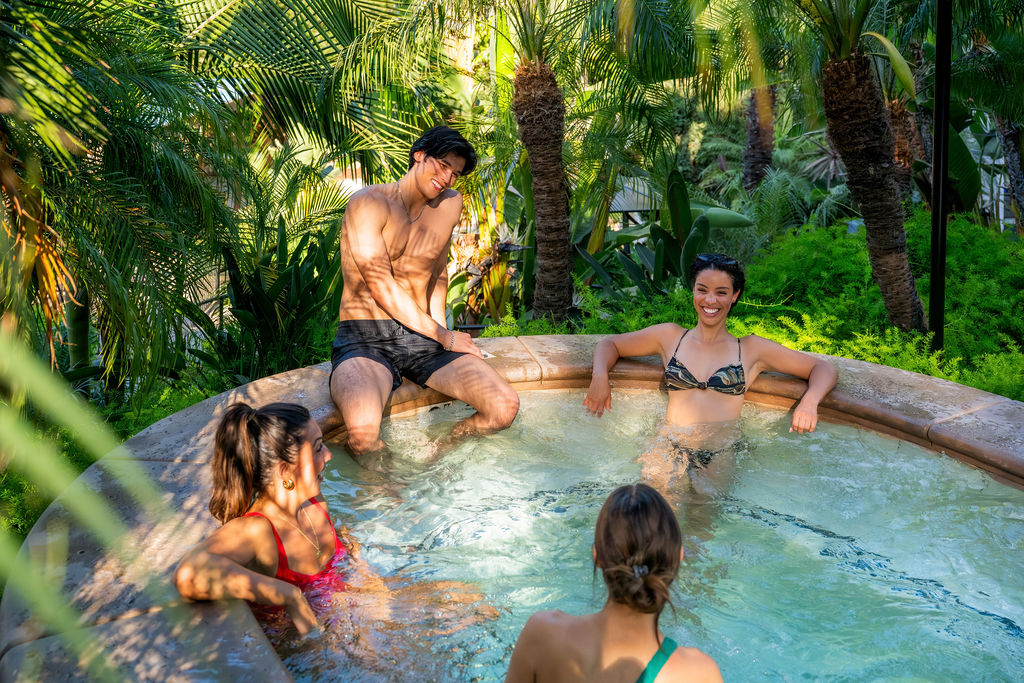 A group of people relax in a tropical outdoor hot tub surrounded by lush palm trees and greenery.