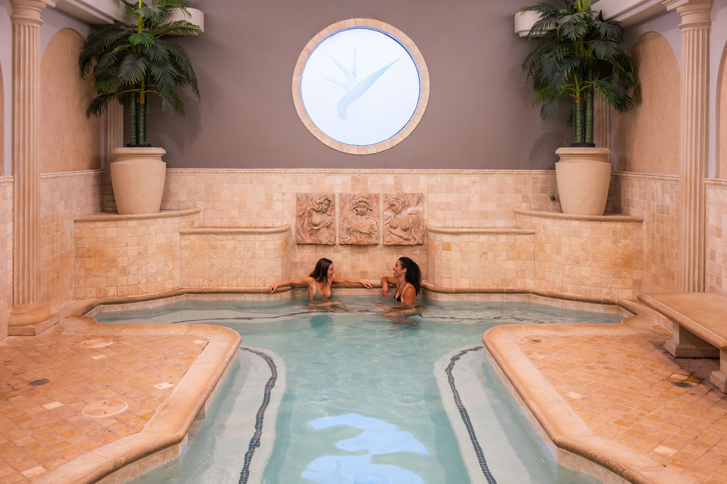 A spa pool with two women in the water, surrounded by palm trees and ornate columns in a warm indoor setting.