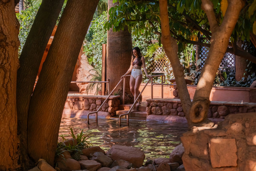 A woman in a white swimsuit stands by a tropical pool, surrounded by trees, rocks, and terracotta walls.
