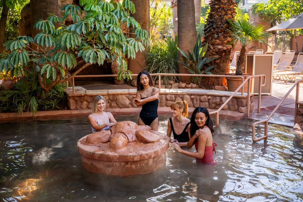 Four women relax in a small outdoor hot tub with bubbles and steaming water beside tropical plants, in a sunny resort setting.