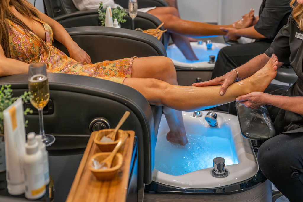 A spa scene with people getting manicures/pedicures and foot-washing in a pedicure chair with trays and champagne nearby.