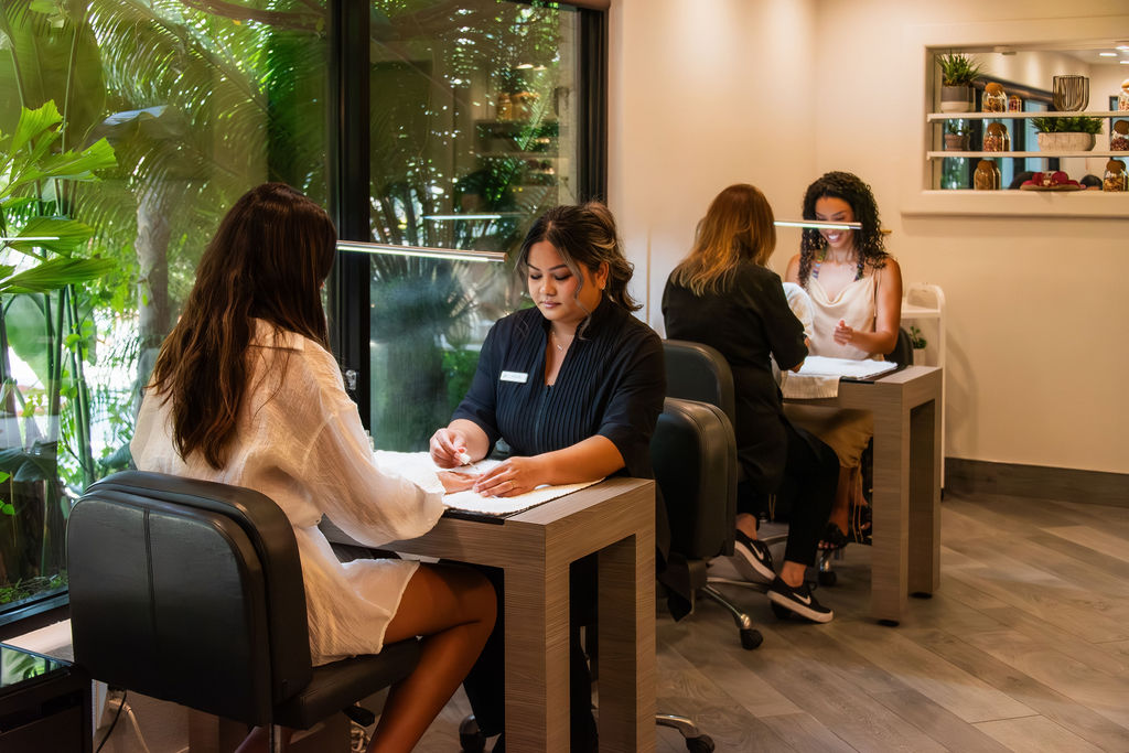 In a bright office, several women sit at desks reviewing documents and chatting with a coworker.