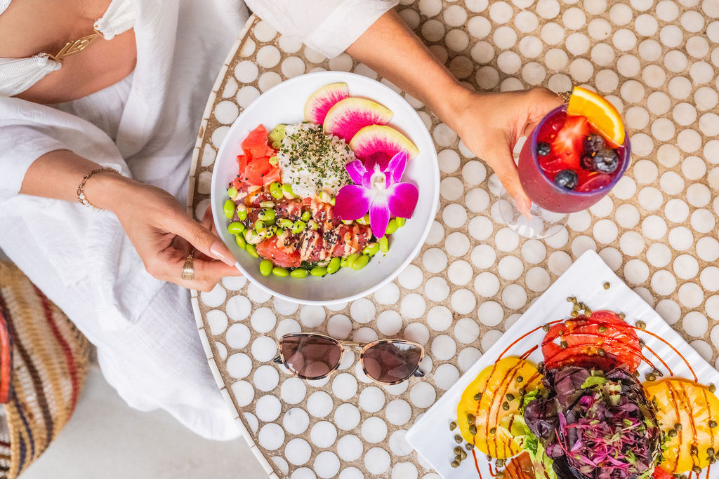 Two people eat a colorful salad on a table with watermelon slices, a berry drink, fruit garnishes, and sunglasses, serving a fresh outdoor meal.