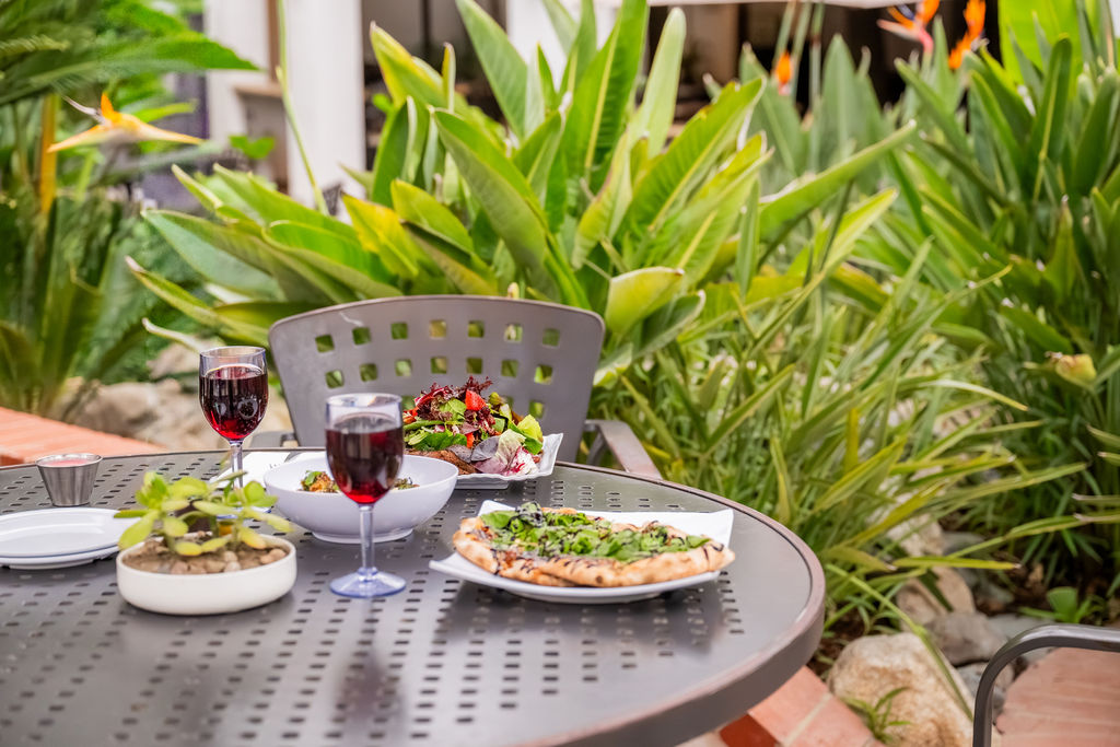 An outdoor patio dining set with plates of food and two glasses of red wine among lush green plants, for a sunny meal.