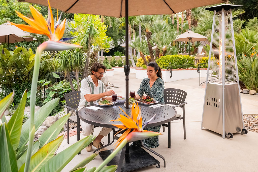 A couple dining at a patio table in a lush tropical garden, with umbrellas and a tall outdoor heater nearby.
