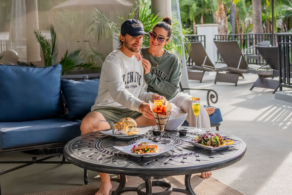 A couple lounges outdoors at a sunny patio table, smiling as they enjoy meals and drinks together beside palm plants.