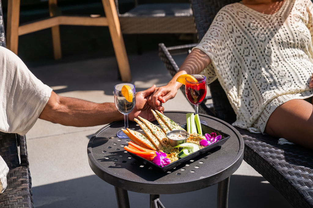 Two people clink wine glasses over a grill tray with vegetables like carrots, asparagus, and purple flowers at an outdoor meal.