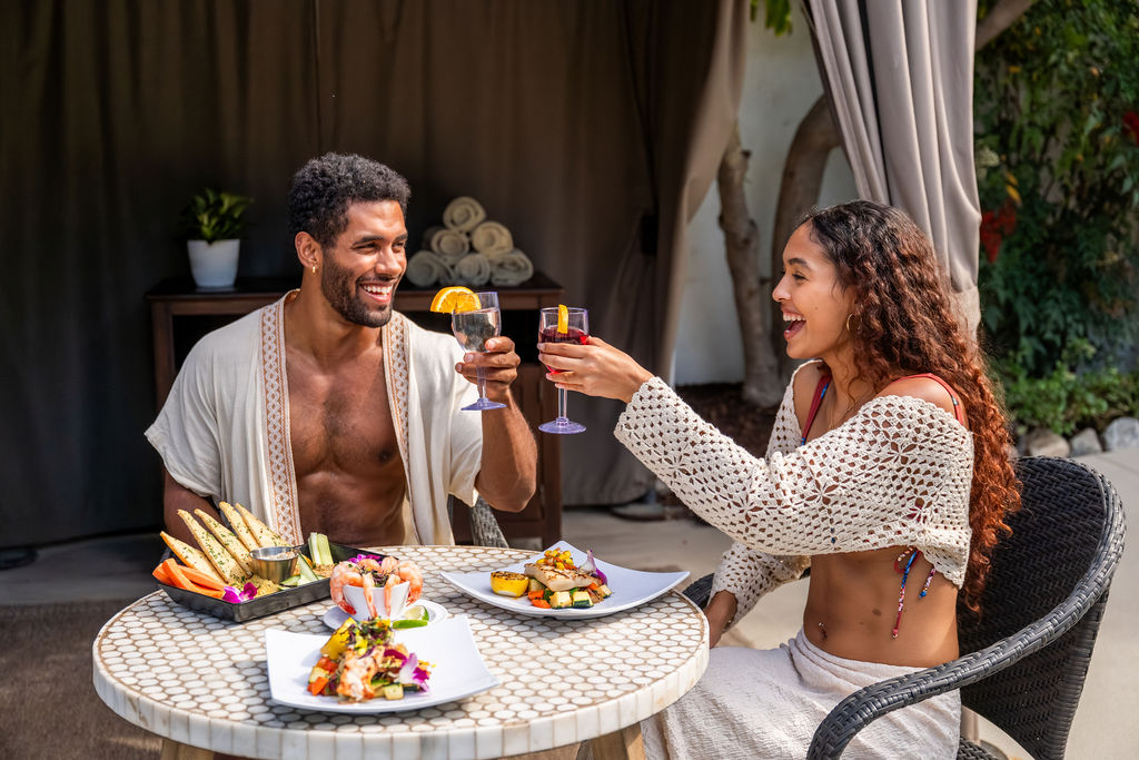 A couple cheers with drinks while enjoying a romantic outdoor meal with colorful plates and appetizers, smiling together.