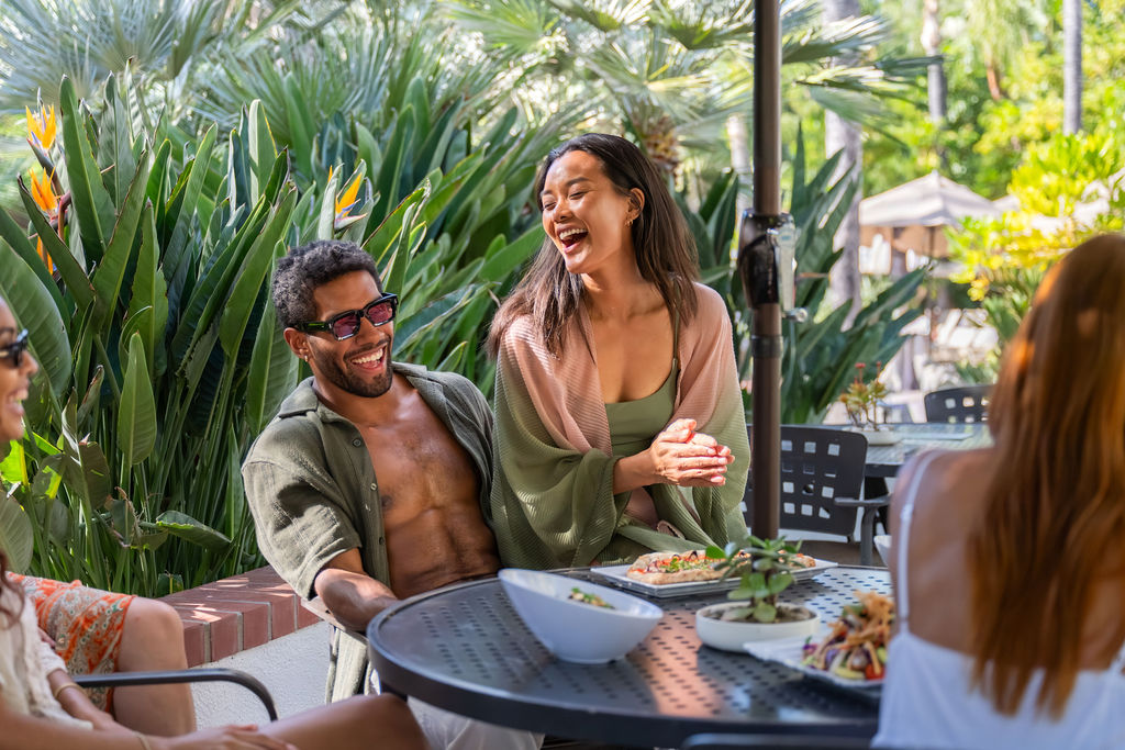 A group of friends enjoy an outdoor meal on a patio, laughing beside a table filled with food and drinks.