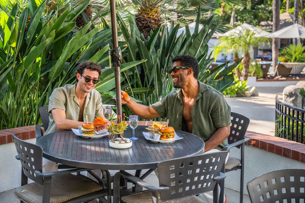 Two friends enjoy a sunny outdoor lunch at a round table with burgers and drinks, surrounded by lush tropical plants.