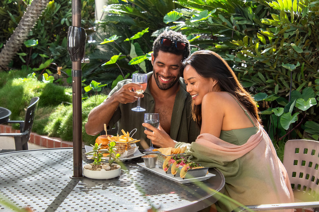 A couple dining outdoors in a lush garden, smiling with drinks and tacos/food on a perforated table in the sunshine, yes.