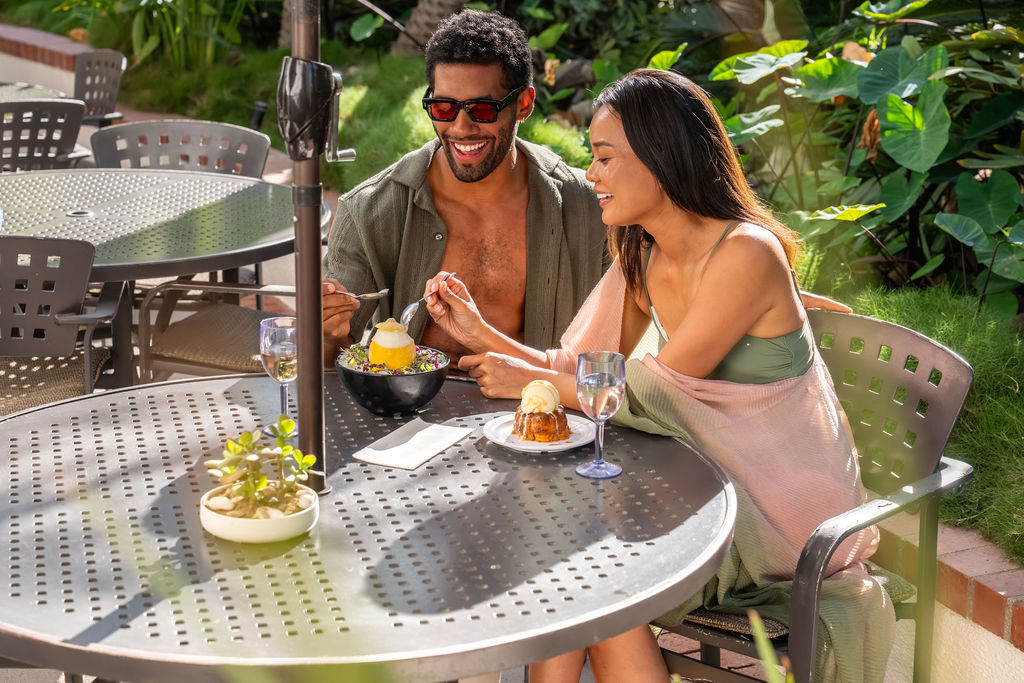A man and woman dine outdoors at a patio table, enjoying desserts and drinks together, smiling in a lush garden setting.