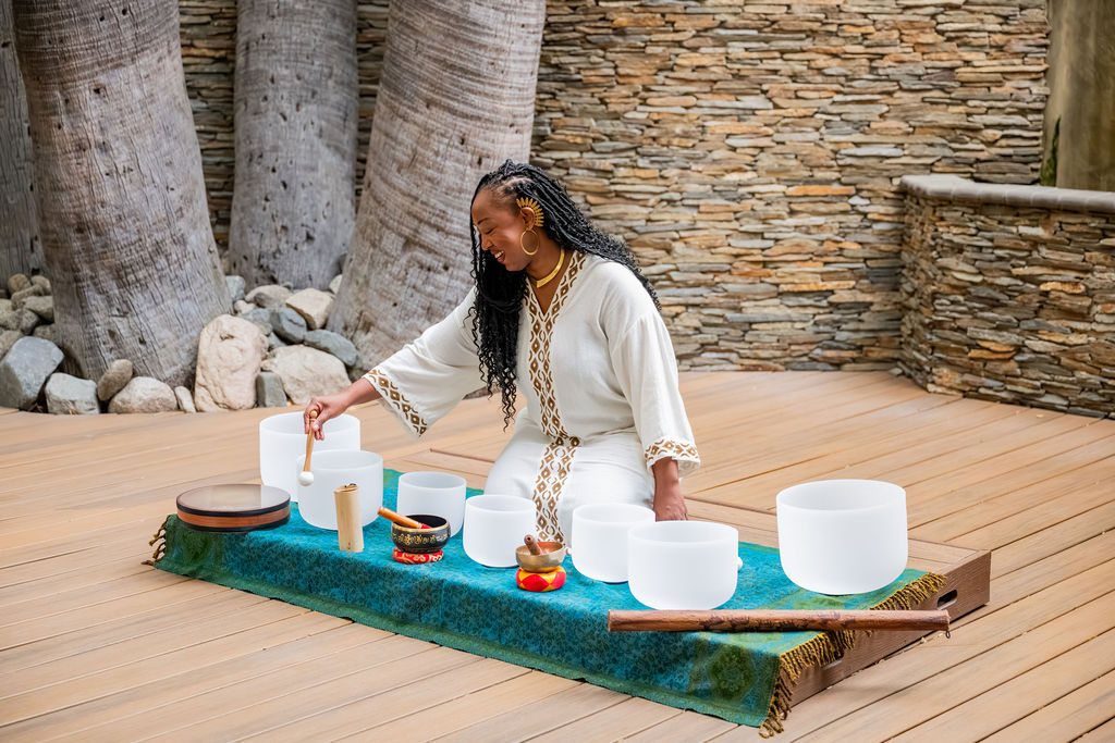 A woman in white traditional attire arranges steaming white tea cups on a small table outdoors, with stone walls behind her, right at a tea service.