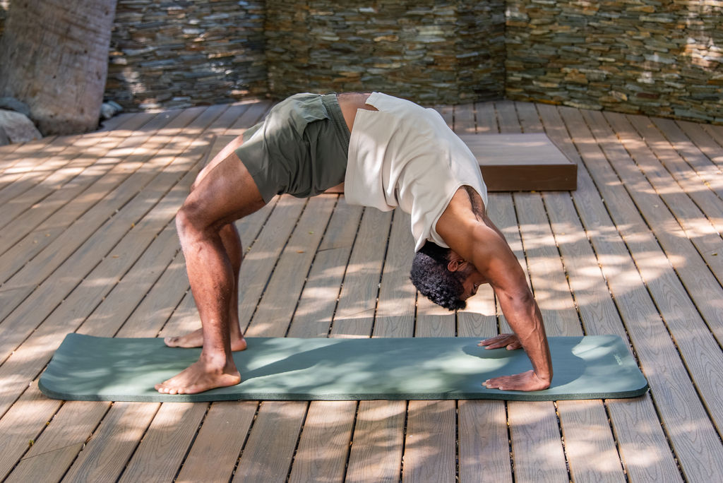 A man doing a yoga plank stretch on a mat outdoors, bending down with hands and feet planted on a wooden deck, ending in a downward-dog pose.