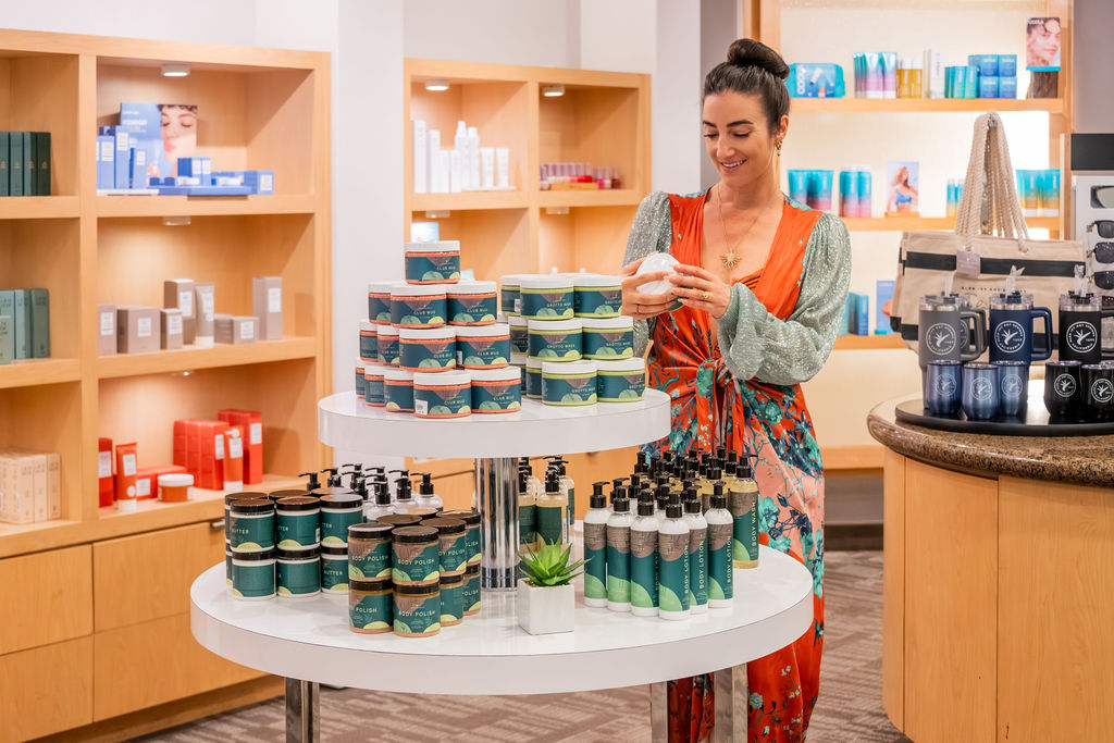 A woman in a beauty shop smiles while arranging skincare items like polish, sprays, and candles on display tables, ready for customers.