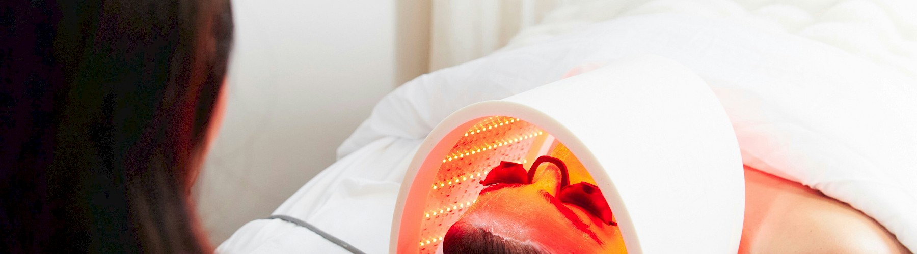 A woman getting a facial under a red LED light lamp in a salon setting, with another person assisting.