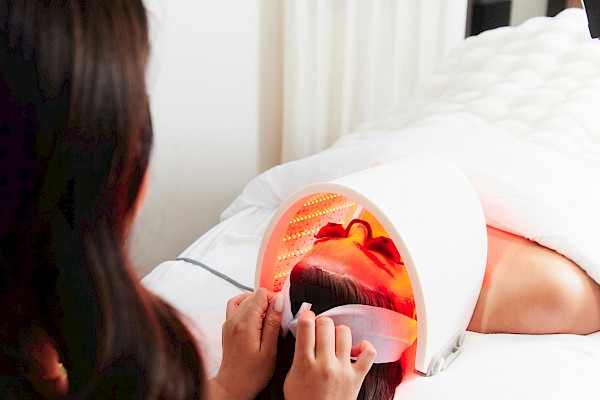 A woman getting a facial under a red LED light lamp in a salon setting, with another person assisting.